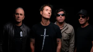 Fotografia promocional da banda de rock brasileira Capital Inicial. Os quatro integrantes estão posicionados lado a lado contra um fundo preto sólido. À esquerda, um integrante usa uma jaqueta de couro preta sobre uma camiseta escura. Ao centro, o vocalista Dinho Ouro Preto veste uma camiseta preta com a estampa de uma espada vertical. À sua direita, o terceiro integrante usa uma jaqueta estilo militar na cor cáqui. Na extremidade direita, o quarto integrante usa óculos escuros, camiseta preta e um boné escuro com o logotipo de uma caveira coroada. Todos mantêm expressões sérias e uma postura clássica de banda de rock.